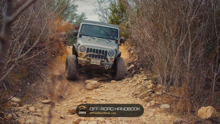 Gray Jeep Wrangler JK crawling down a rocky, brush-lined dirt trail toward the camera