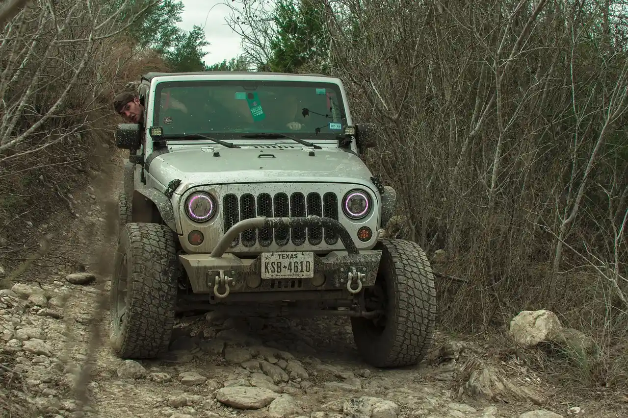 Gray Jeep Wrangler with a front bumper and all-terrain tires crawling over a rocky, brushy trail