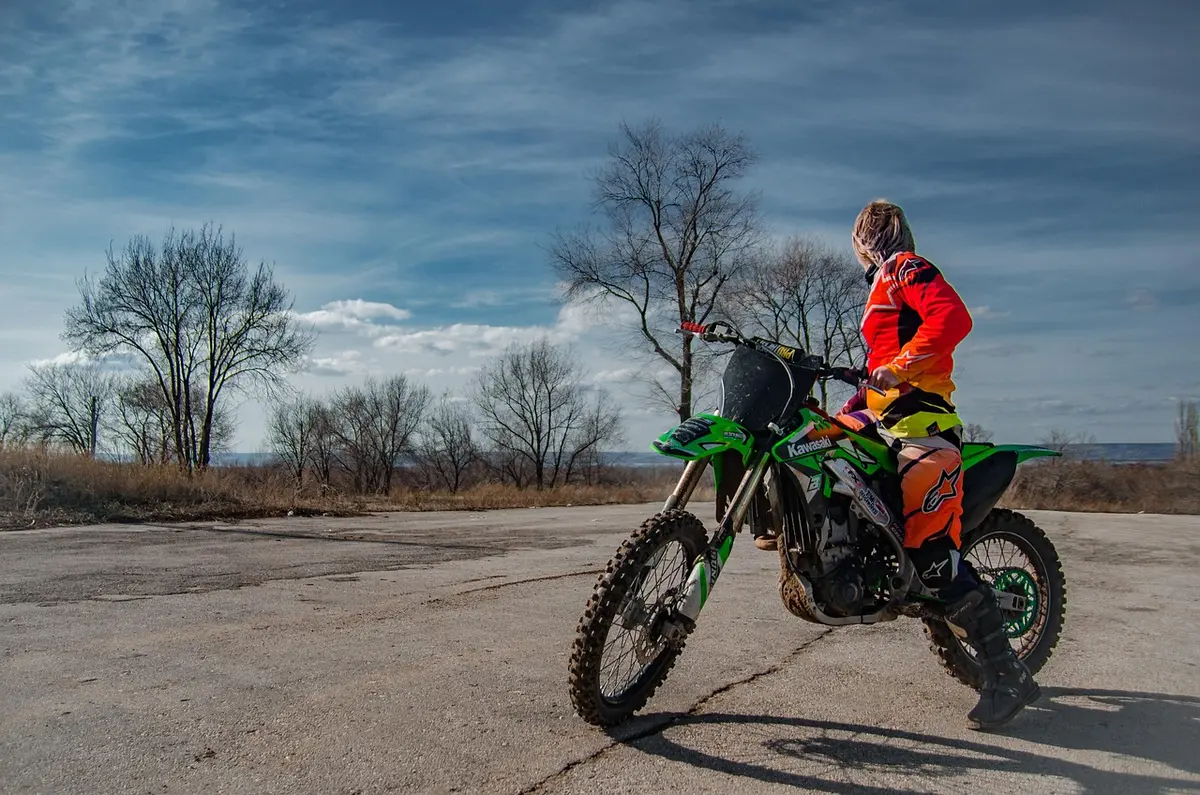 Rider in orange gear sitting on a green Kawasaki motocross bike on a paved lot