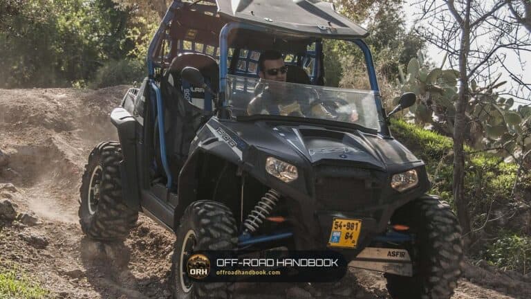 Dark side-by-side UTV climbing a dirt trail slope past brush with the driver at the wheel