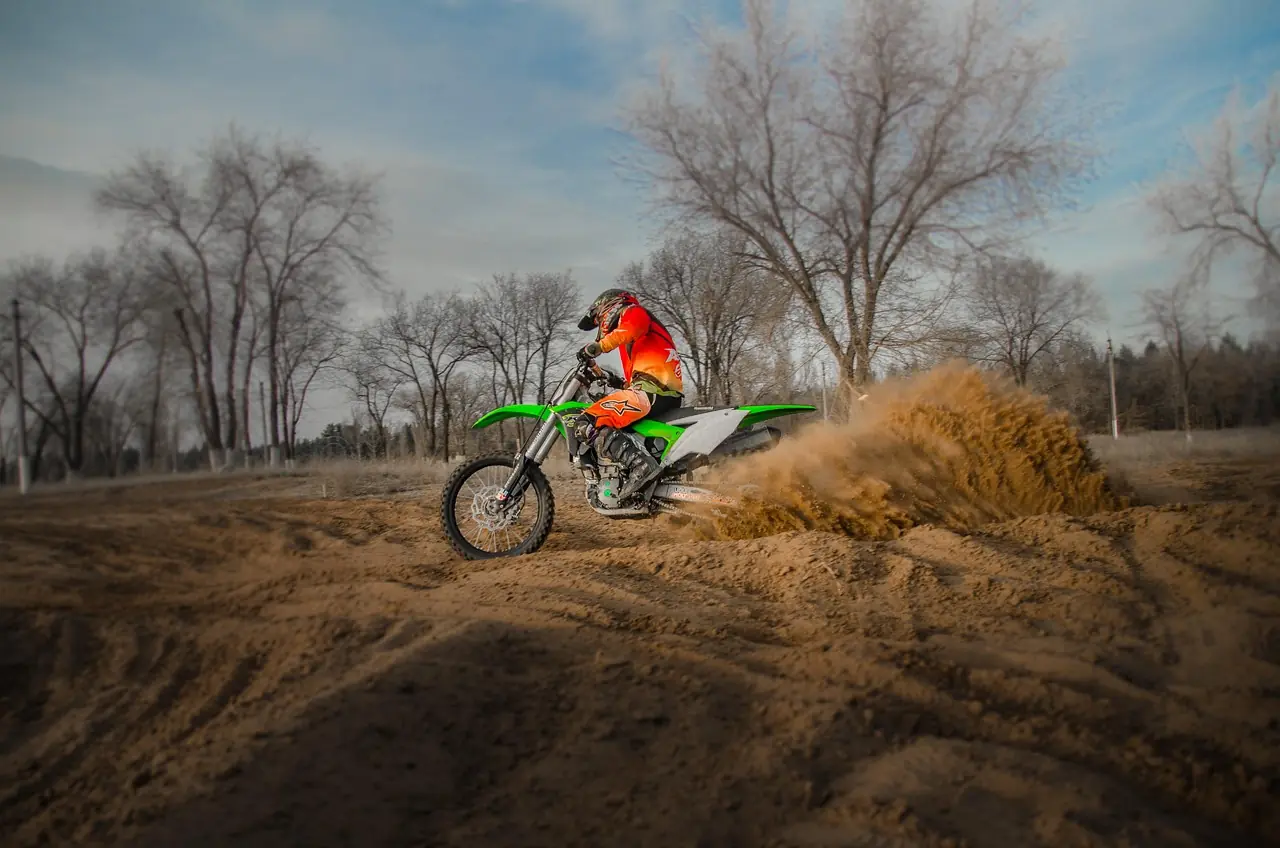 Rider in orange gear on a green Kawasaki motocross bike throwing a roost of dirt out of a sand turn