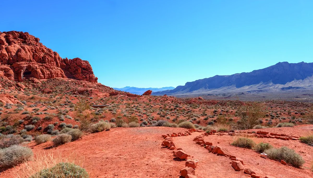 A red-sand desert trail with red rock outcrops and blue mountains in the distance near Las Vegas