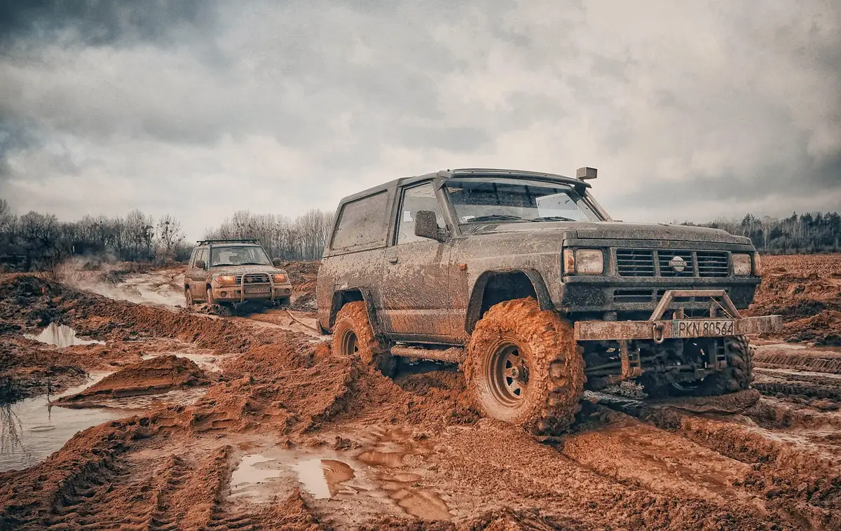 SUV with a mud-covered body driving through deep red-brown ruts on an off-road trail