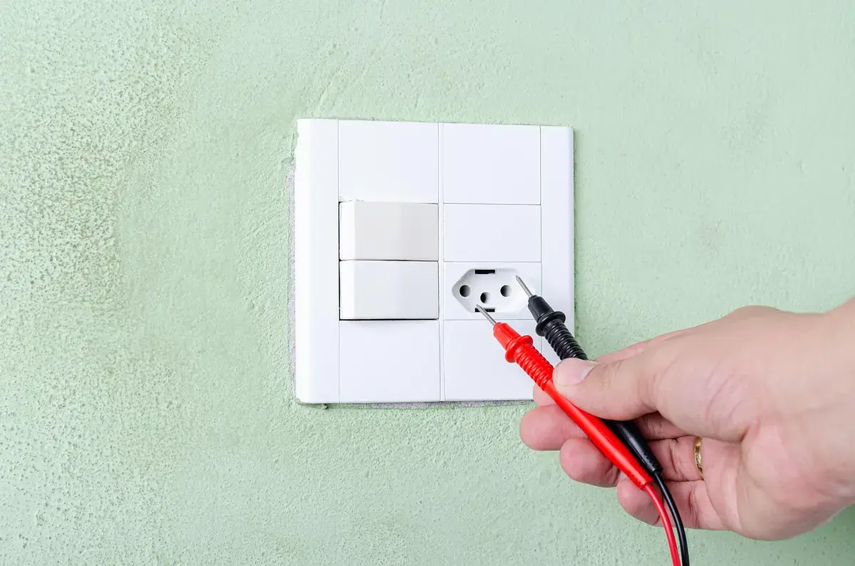 Yellow digital multimeter held with red and black test leads against a wall outlet
