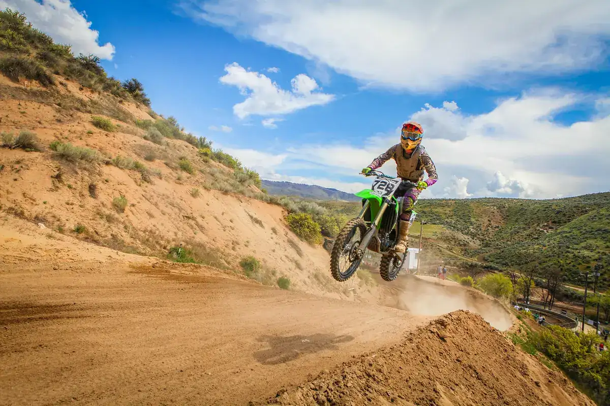Rider on a green motocross bike jumping off a dirt hillside under a blue sky