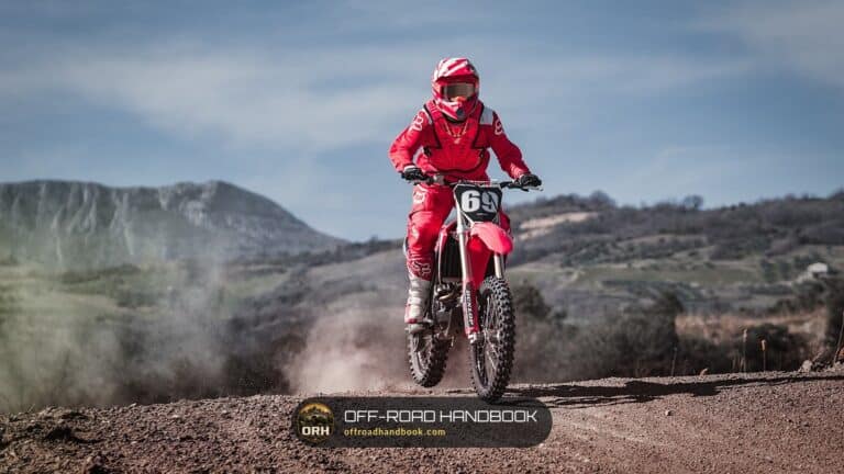 Rider in red gear on a red motocross bike kicking up dust on a dirt track with hills behind