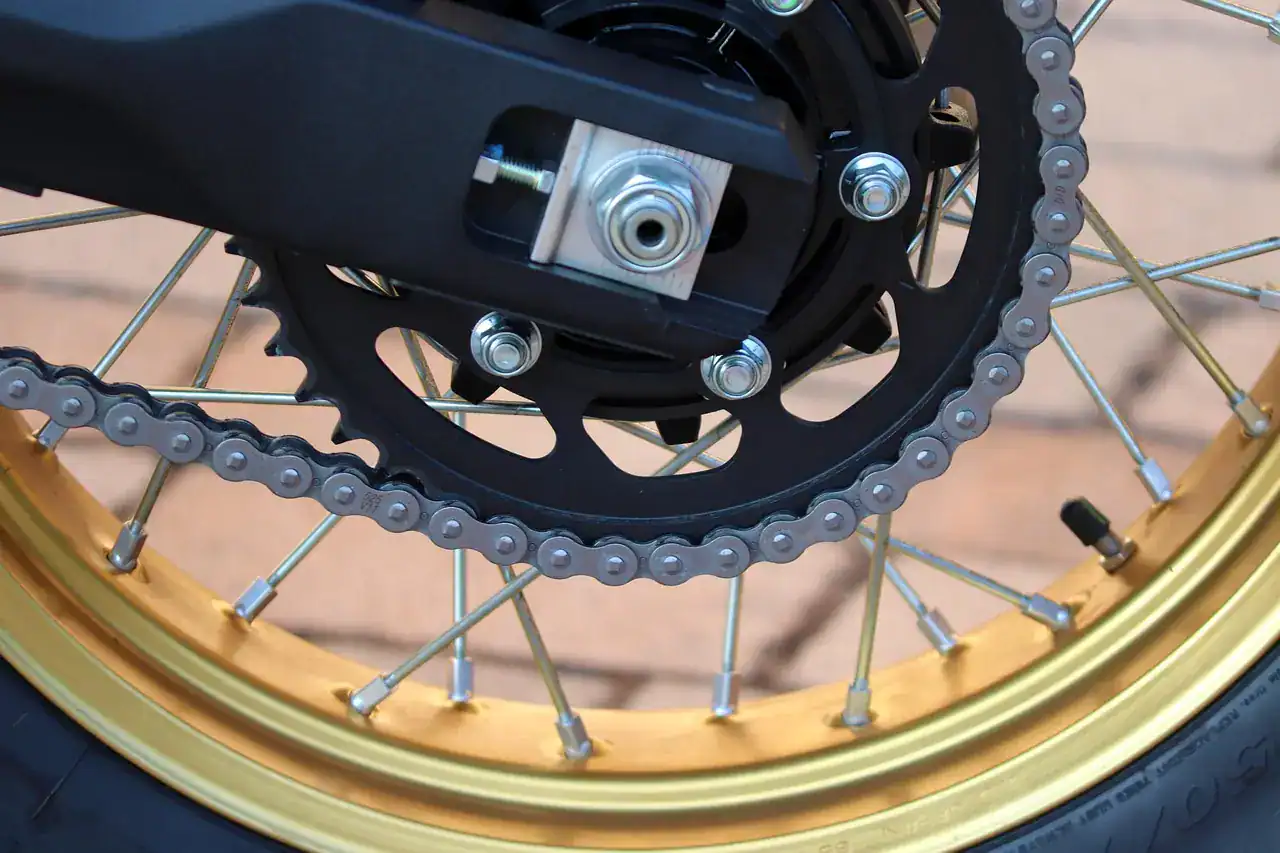 Black rear sprocket with chain and axle adjuster on a gold spoked motorcycle wheel, shown to illustrate final drive