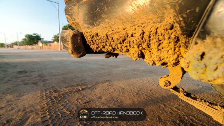 Close-up of a muddy 4x4 front end and recovery hook sitting low over a dirt road, showing how little clearance sits under the bumper