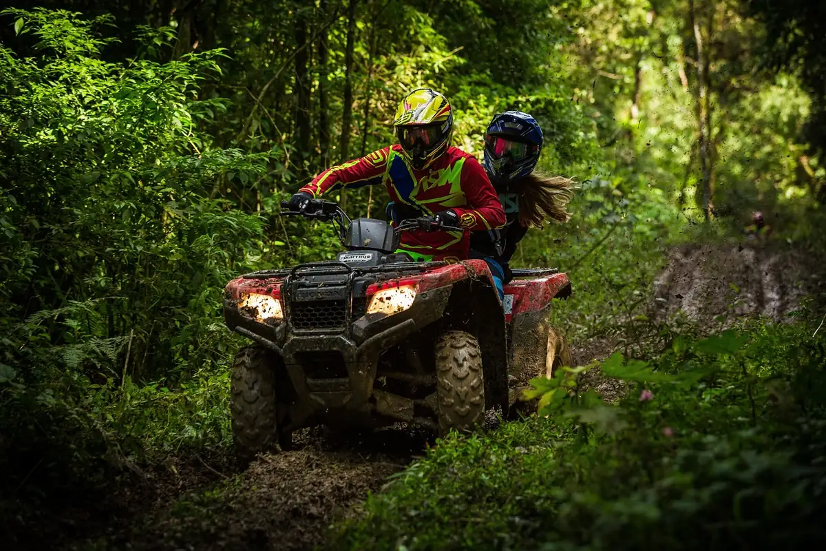Muddy utility ATV with two riders on a dark forest trail