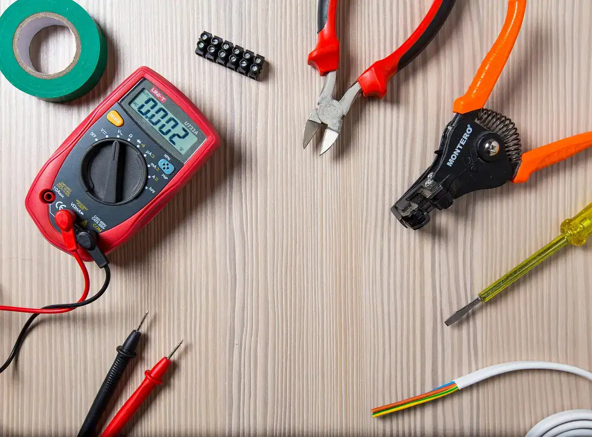 Digital multimeter with red and black test leads, wire cutters, wire stripper and a screwdriver on a wooden bench