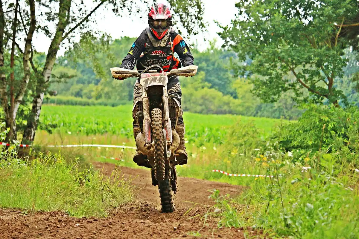 Dirt bike rider in a helmet and goggles on a muddy dirt trail between grass and trees