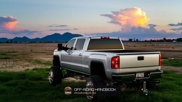 Rear three-quarter view of a lifted silver pickup truck with large aftermarket off-road tires parked in a field at sunset