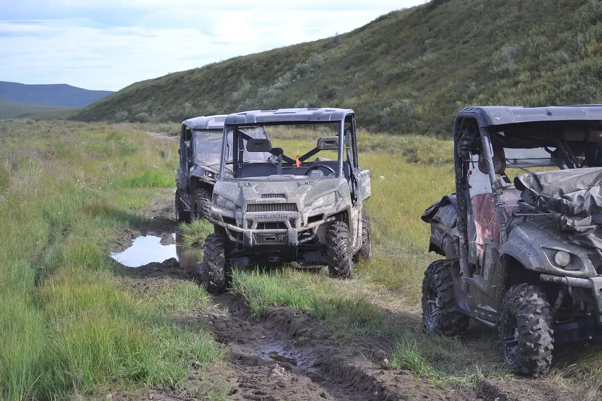 Two muddy side-by-side UTVs on a wet grassy trail below a hillside