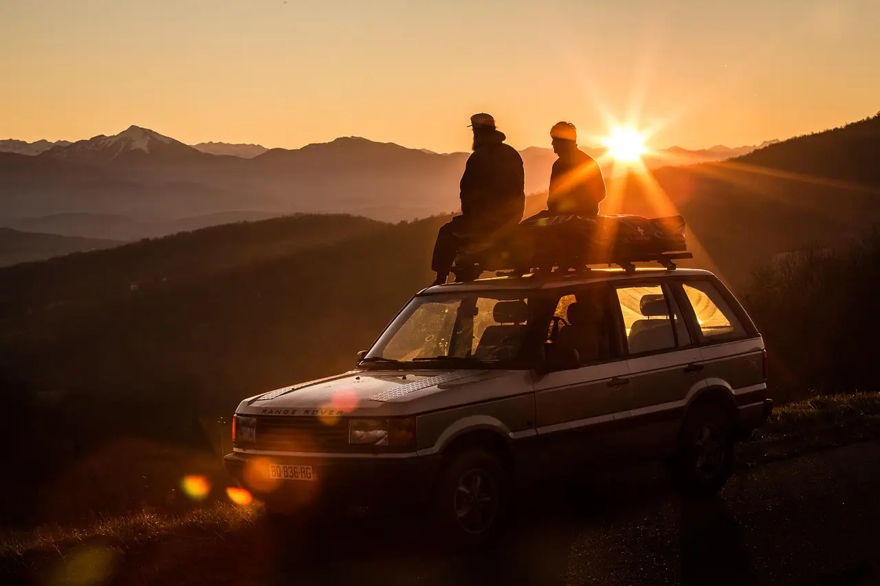 Older wagon with two people sitting on the roof rack watching the sun set over mountains; a generic overland vehicle