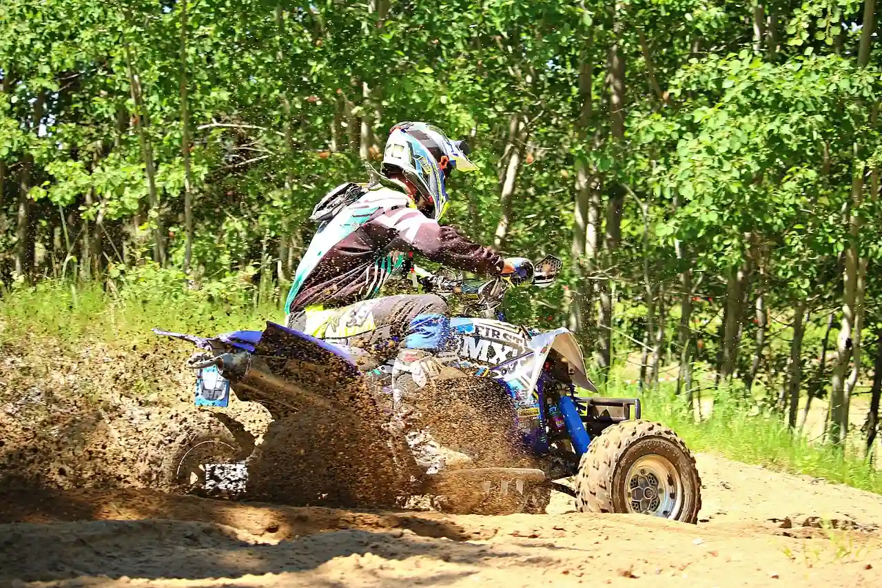 Rider in a helmet and riding gear on a sport ATV kicking up dirt on a sandy trail beside trees