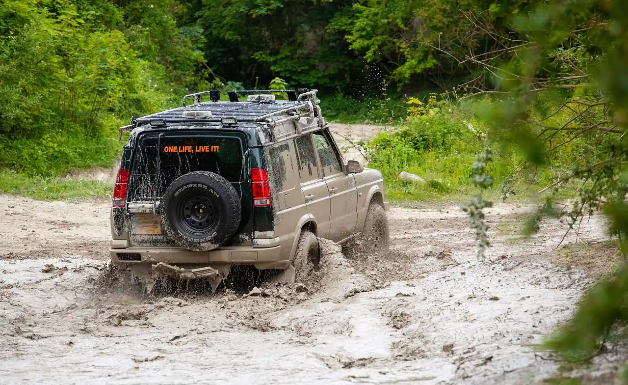 SUV with a roof rack and spare tire driving through a wet, muddy sand trail beside trees