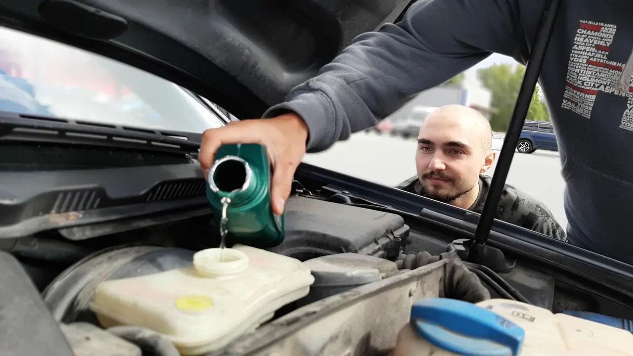 Person pouring fluid from a green bottle into a plastic reservoir under a raised car hood
