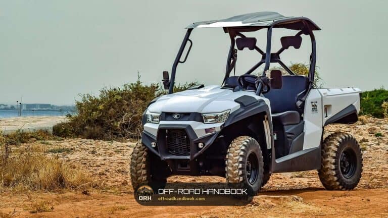 White side-by-side utility vehicle with a roof parked on a sandy dirt trail beside scrub brush