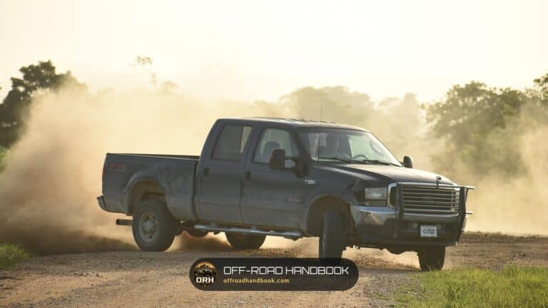 Dark crew-cab pickup truck driving on a gravel road with dust hanging in the air behind it