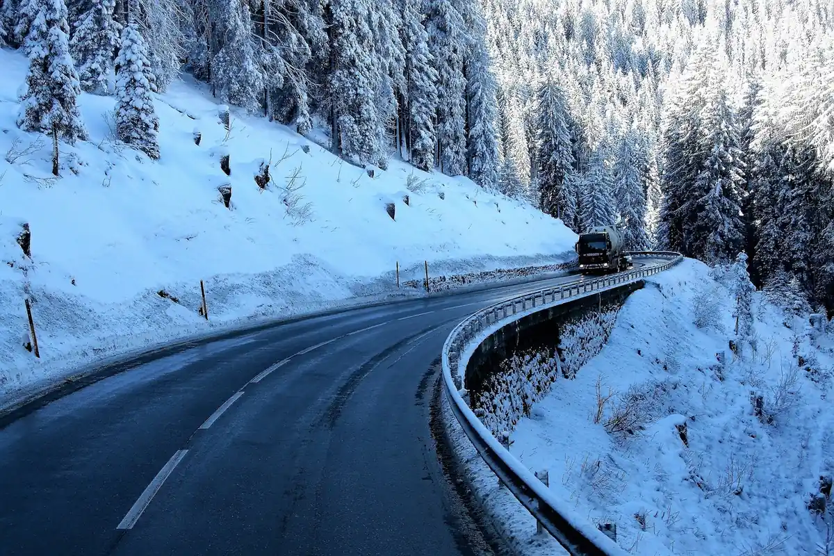 Plowed, wet two-lane mountain highway curving through snow-covered trees with a truck ahead