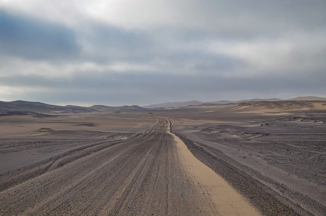 Wide loose-gravel track with tire ruts running across open desert under a cloudy sky