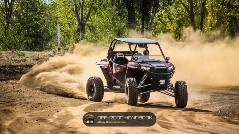 Red and black Polaris RZR sport side-by-side sliding through a dust cloud on a dirt trail in the woods