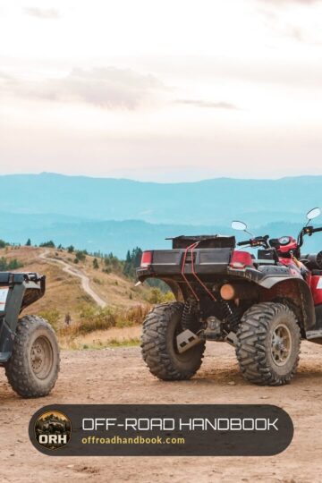 Three utility ATVs, a red one in the center, parked on a dirt overlook with mountains behind