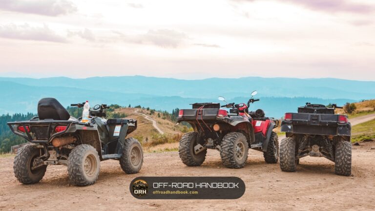 Three utility ATVs, a red one in the center, parked on a dirt overlook with mountains behind