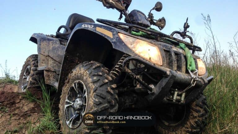 Mud-splattered utility ATV with headlight on climbing a grassy bank, seen from a low angle