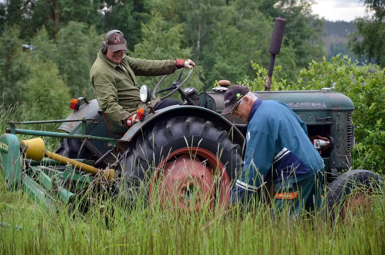 Two men servicing an older tractor in tall grass, one seated at the wheel and one bending over the engine side