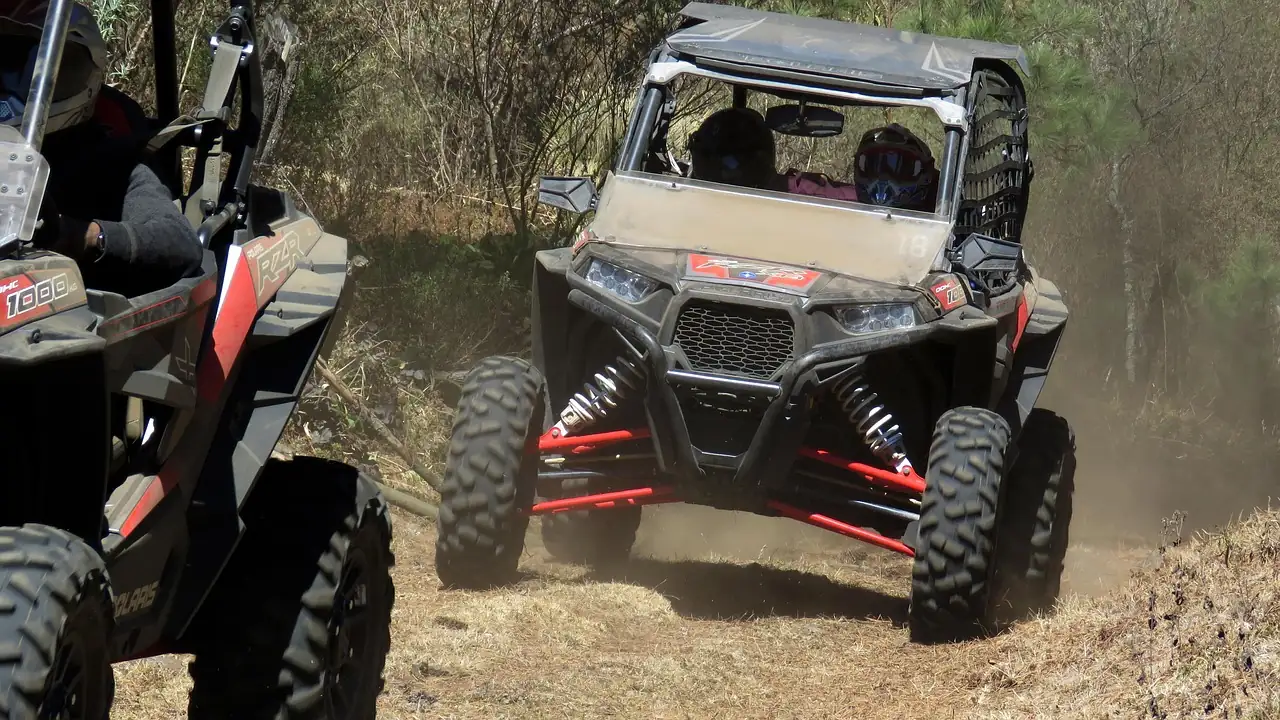 Two Polaris RZR side-by-sides on a dusty dirt trail through dry brush