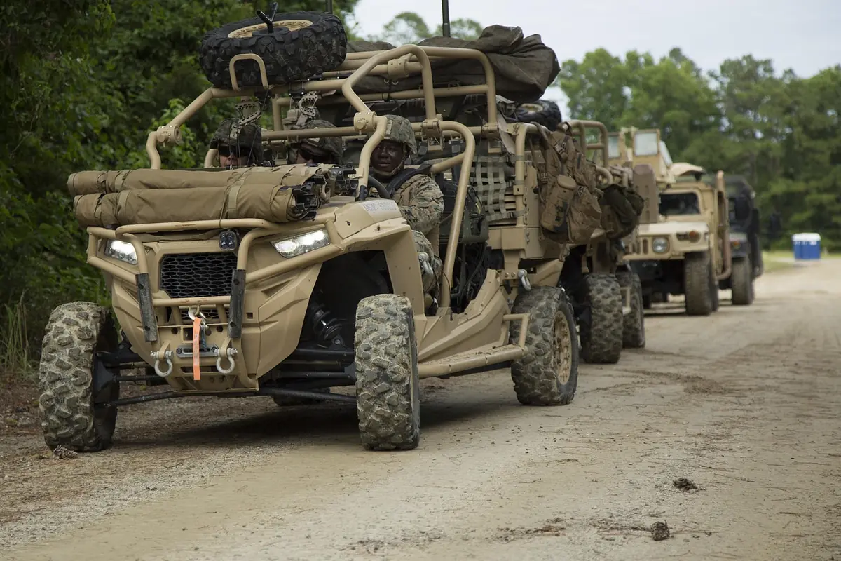 A tan side-by-side with a spare tire and gear strapped on top parked on a gravel road, the kind of loaded low-speed work that is hard on a CVT belt (stock photo of a military-style UTV, not a Pro XP)