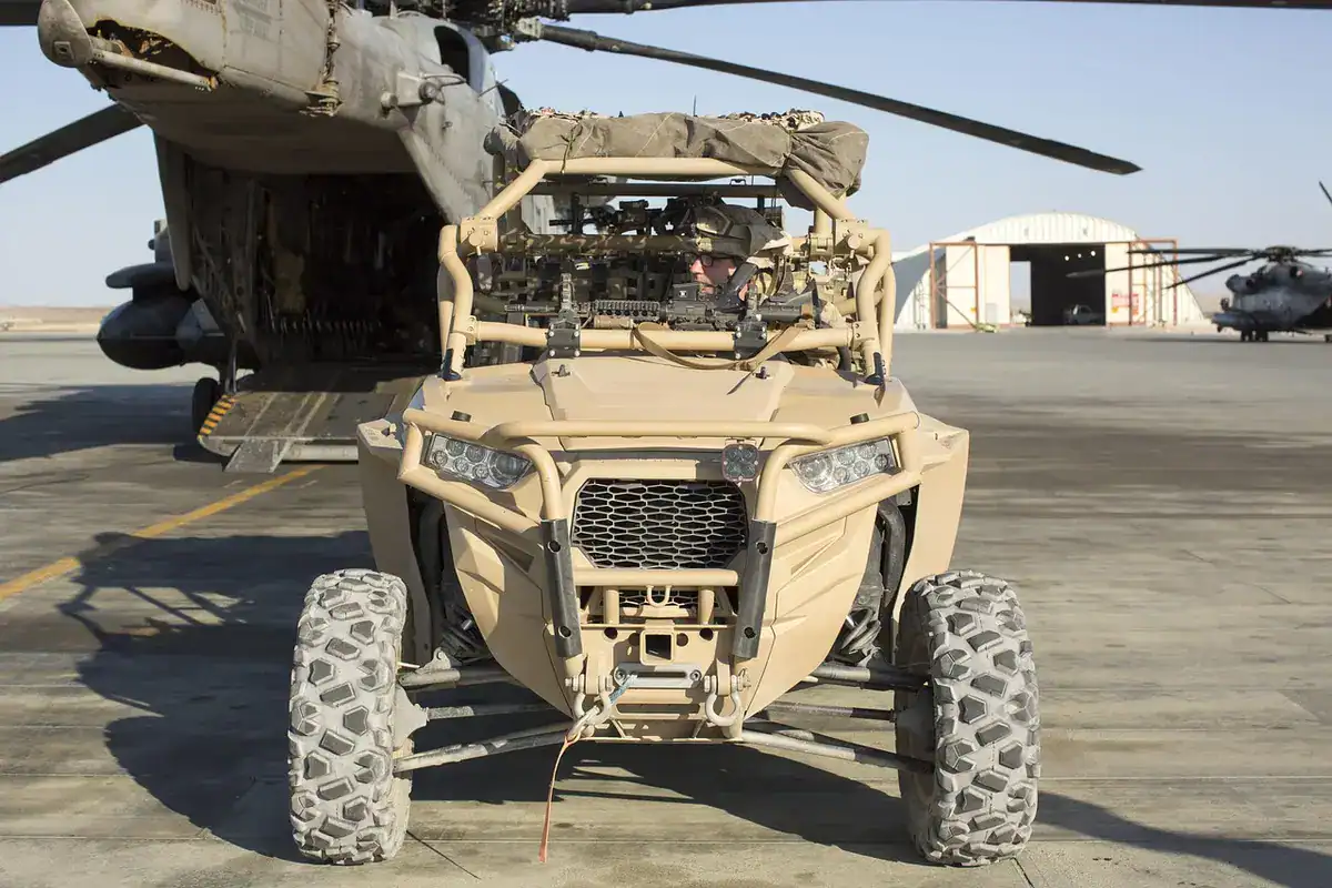 Military-configured side-by-side utility vehicle with roll cage, winch and knobby tires parked beside a helicopter