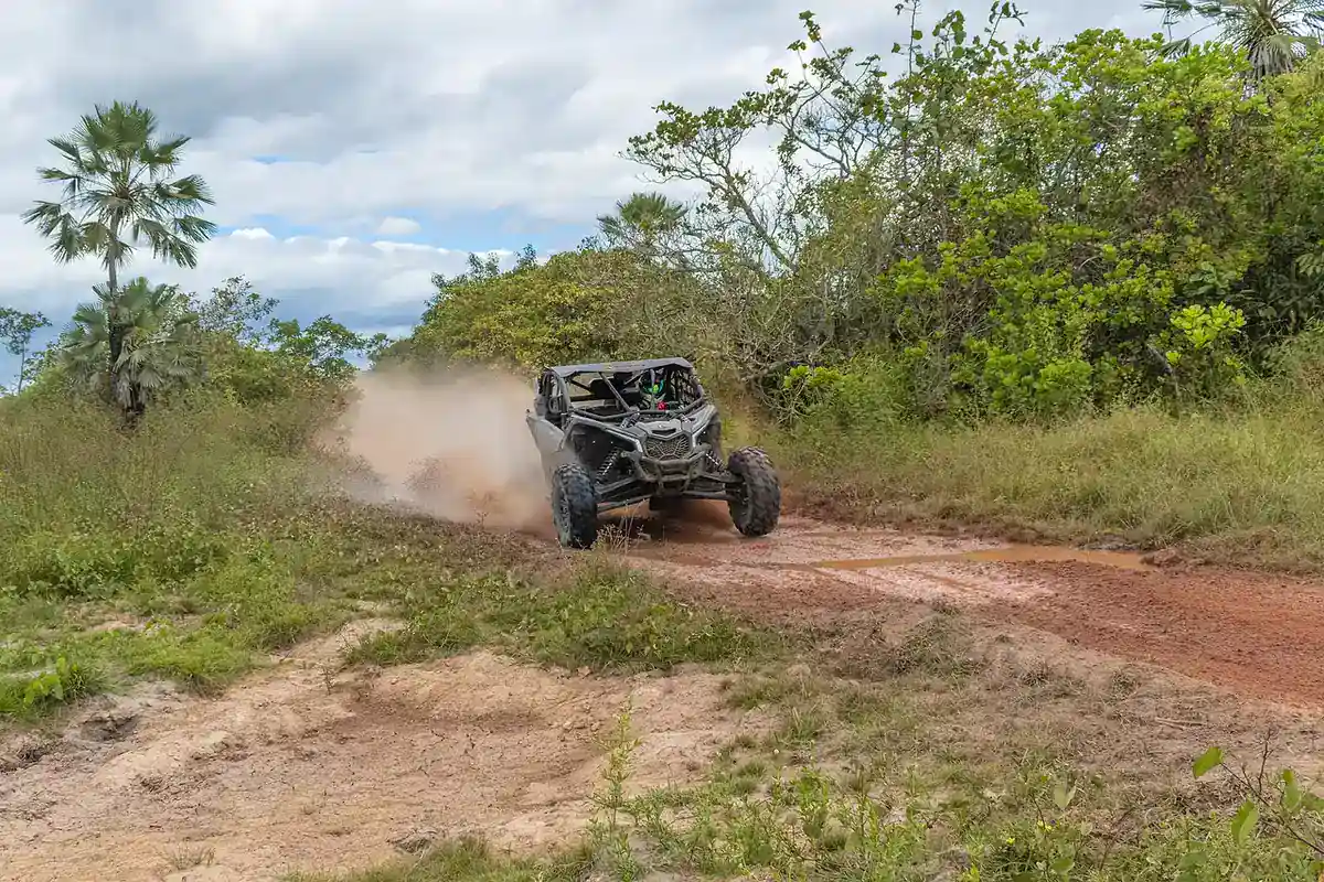 Side-by-side UTV kicking up dust on a muddy red dirt trail through green brush