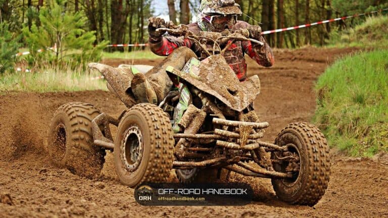 Rider on a mud-covered two-wheel-drive sport quad cornering on a dirt track with trees behind