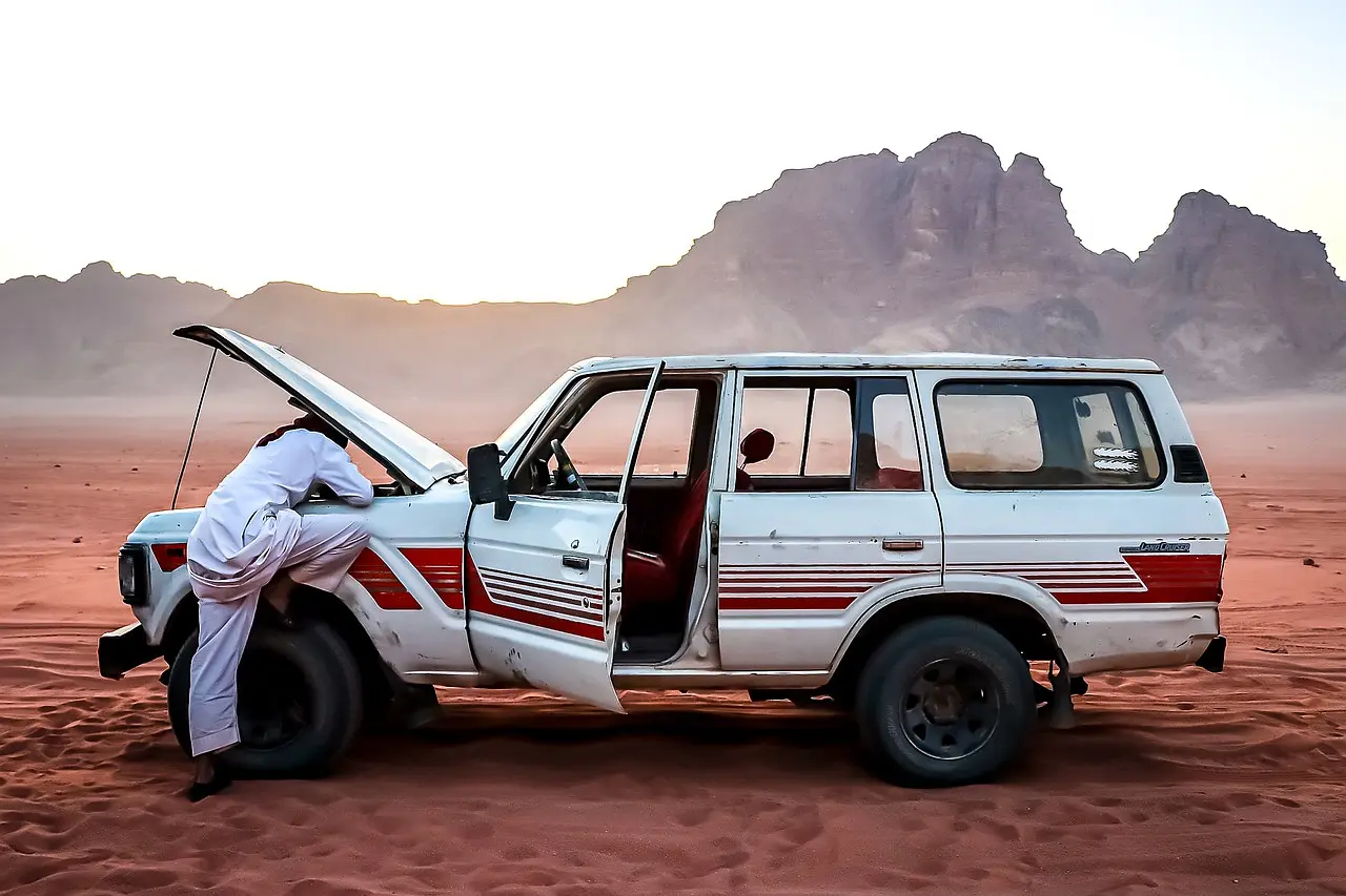 White SUV stopped on a desert road with its hood raised and a man leaning over the engine