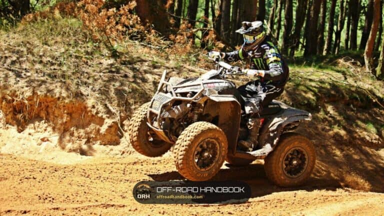 An ATV rider in a helmet crossing a muddy dirt bank beside a pine forest