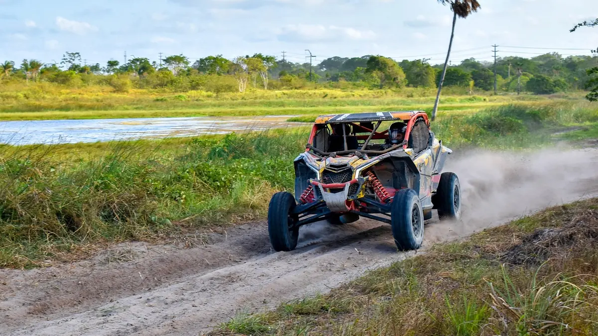 A side-by-side UTV driving fast on a dusty dirt trail with grass and a pond behind it