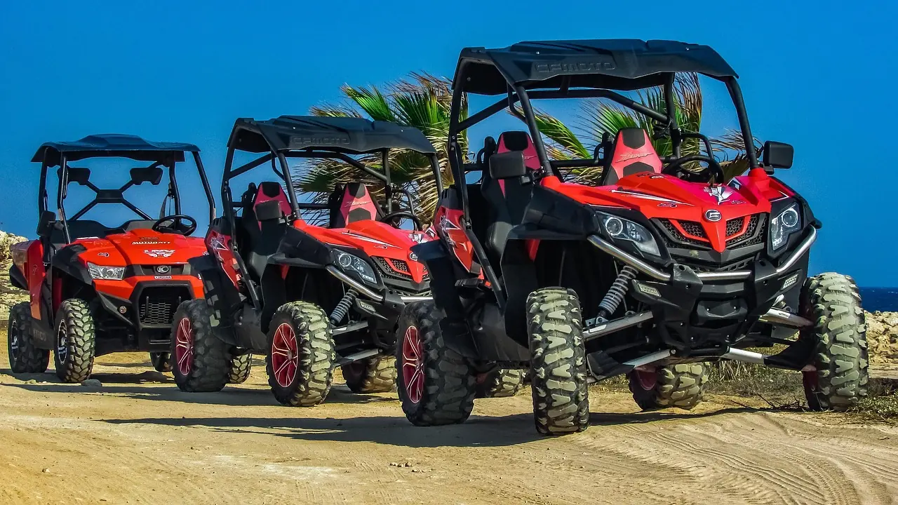 Three red side-by-side UTVs parked in a row on sand beside palm trees