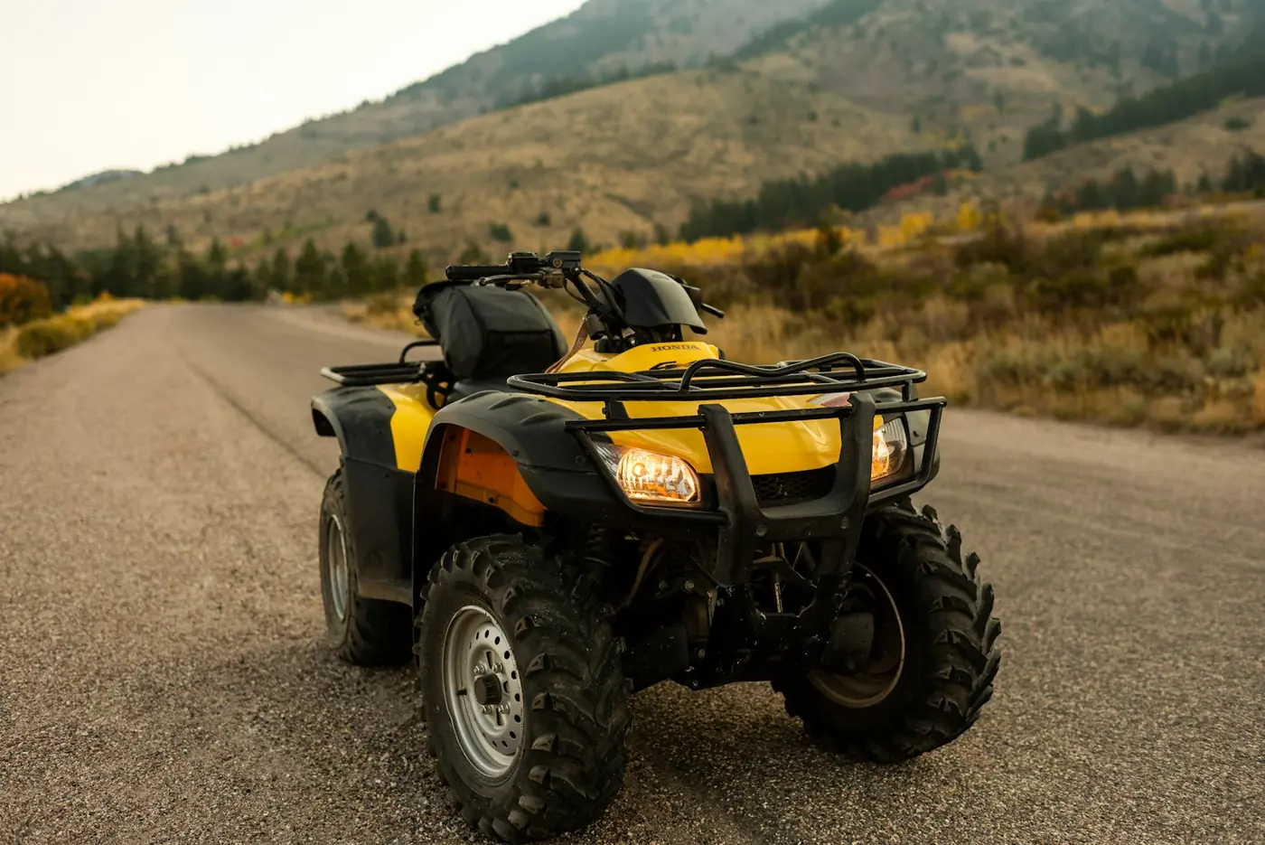 Yellow Honda utility ATV with front and rear racks and headlights on, parked on a gravel road